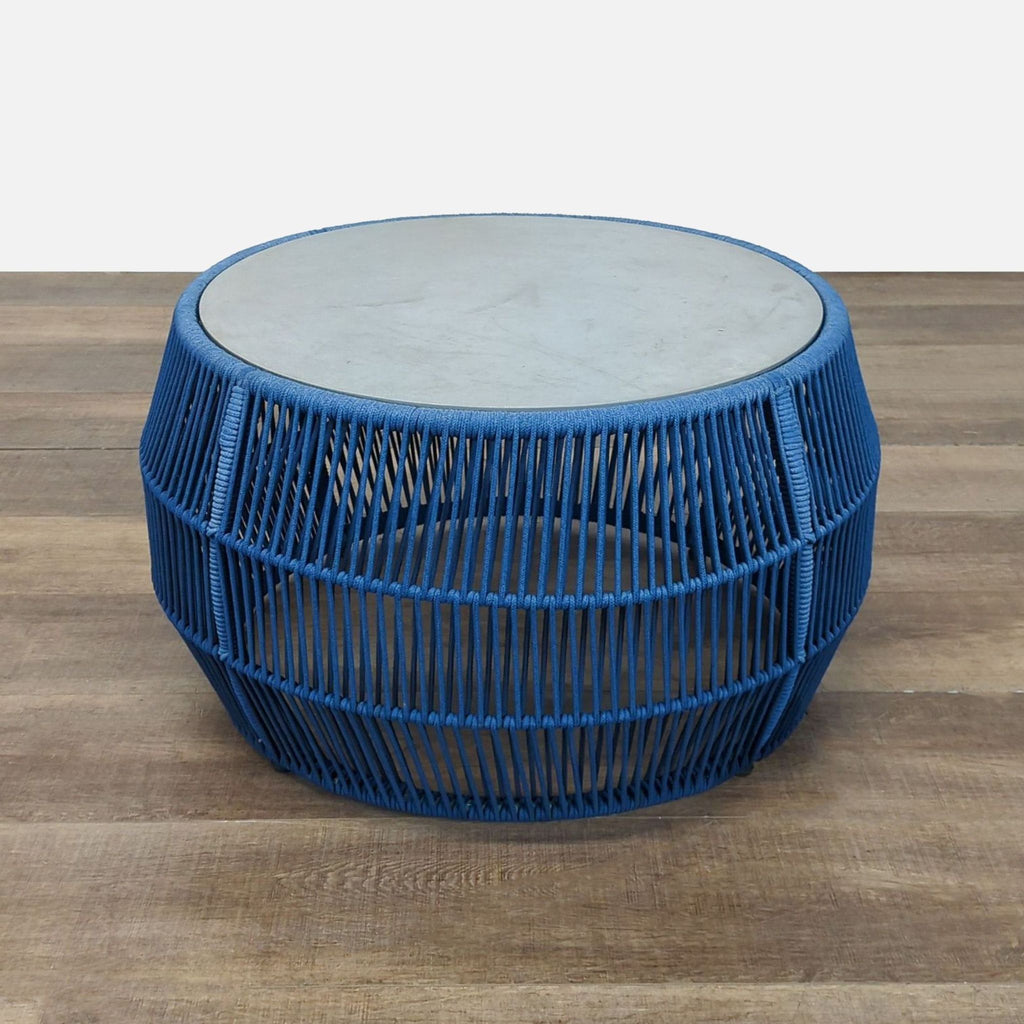Blue rope-woven table by Reperch with a broad base and round stone top, shown on wood floor.  