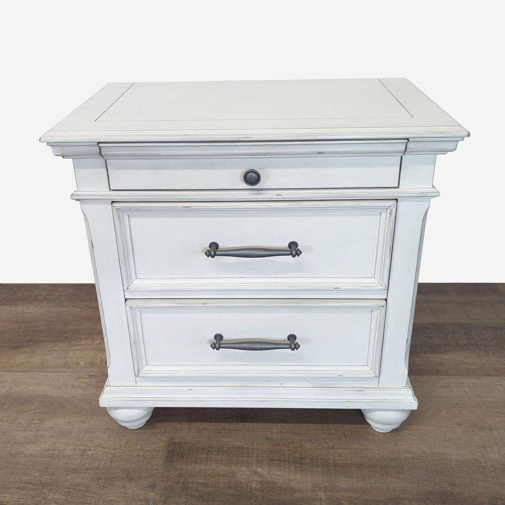 Distressed white Ashley Furniture end table with two drawers and round feet, shown from the front on wood floor.  
