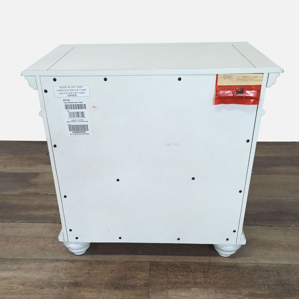 Back view of white distressed Ashley Furniture end table with visible hardware, stickers, and a red attachment.