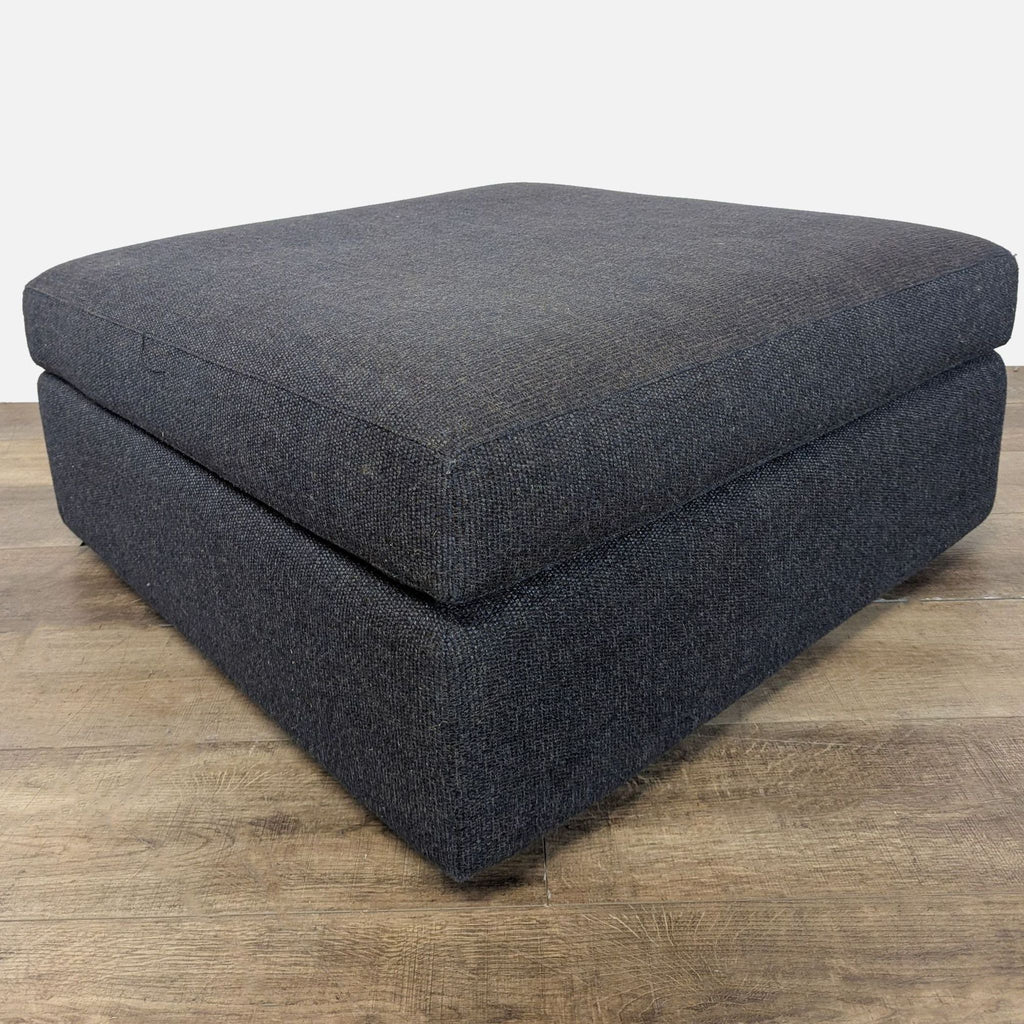 1. Square dark gray upholstered ottoman by Reperch on wooden floor, viewed from a low angle.