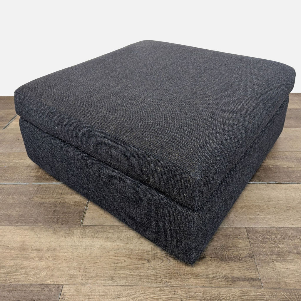 3. Reperch ottoman in charcoal fabric, cube design, placed on wood flooring, shown from the side.