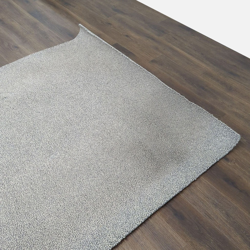 1. Close-up of a textured gray Room & Board rug on dark wood flooring, shown with one corner folded.  