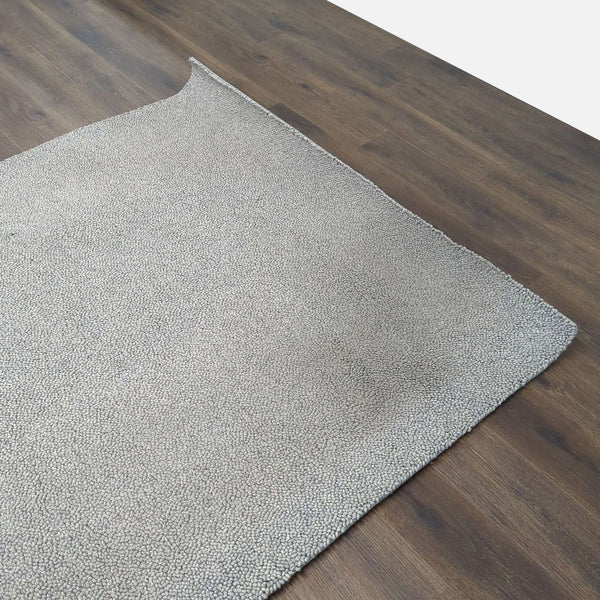 1. Close-up of a textured gray Room & Board rug on dark wood flooring, shown with one corner folded.  