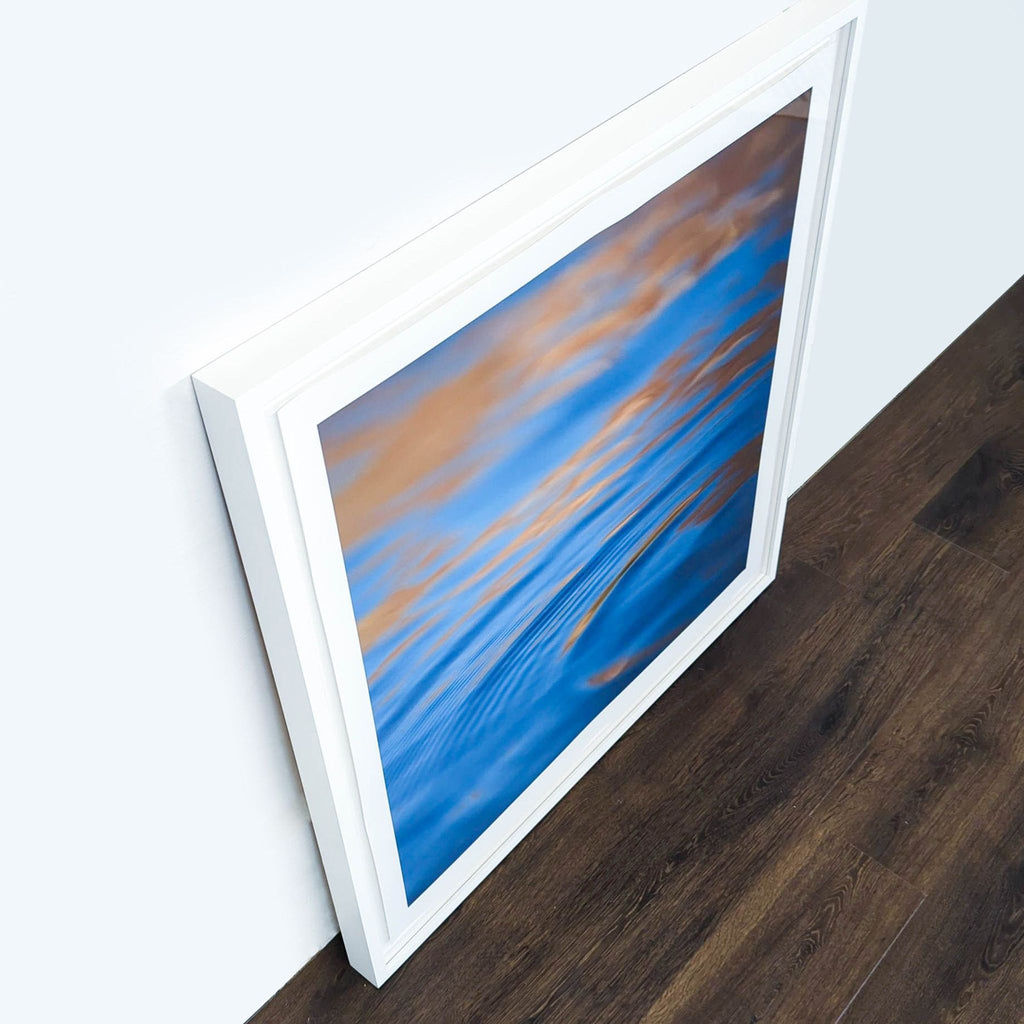 3. Angled view of Reperch abstract water print in a white frame, placed against a wall and dark wood floor.