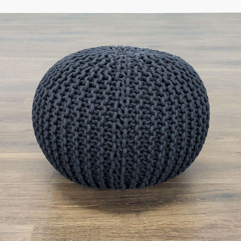 Side view of a Reperch black knitted pouf ottoman, round and textured, set against a wooden floor.  