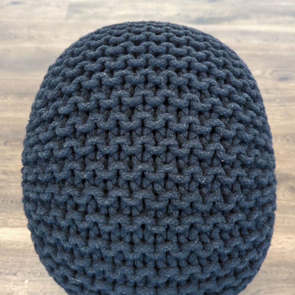 Close-up view of a round, dark knitted pouf with chunky texture from Reperch on a wooden floor.  