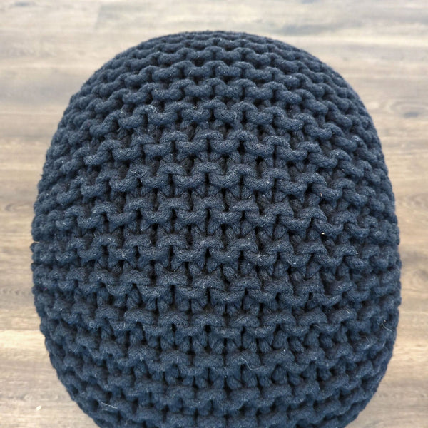 Close-up view of a round, dark knitted pouf with chunky texture from Reperch on a wooden floor.  