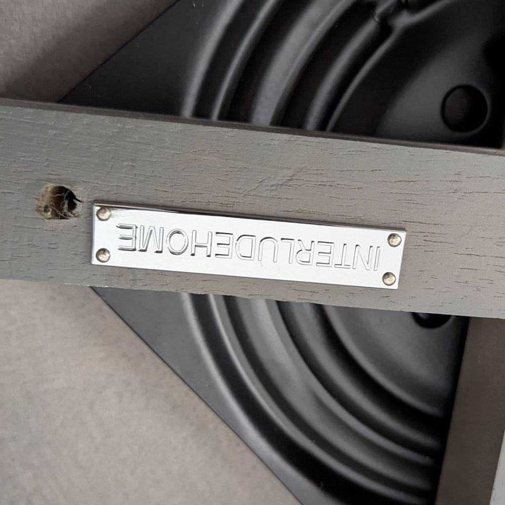 Metal nameplate with "INTERLUDEHOME" branding attached to wooden frame, part of a lounge furniture piece.