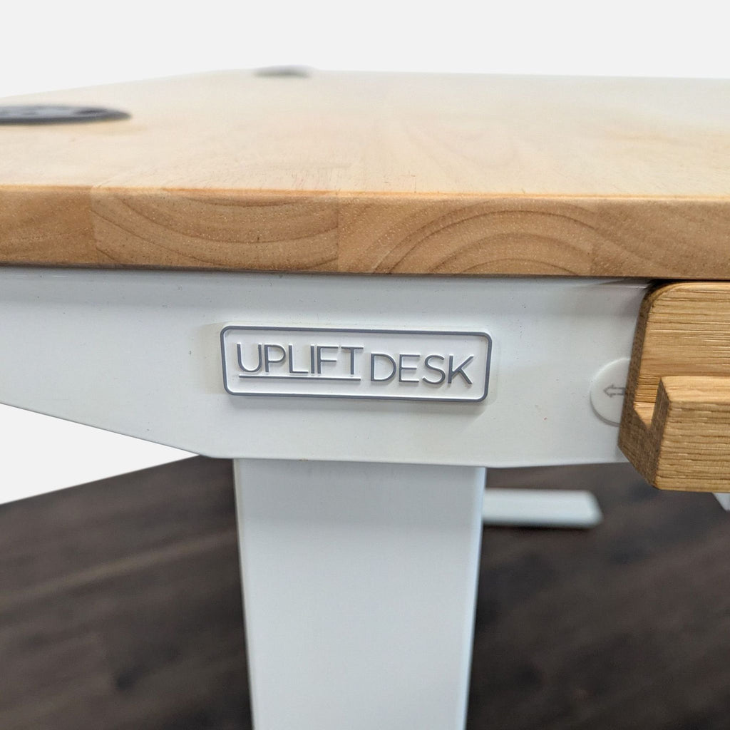 3. Close-up of Uplift Desk branding on a white metal frame under a light wood desktop in the Desks category.