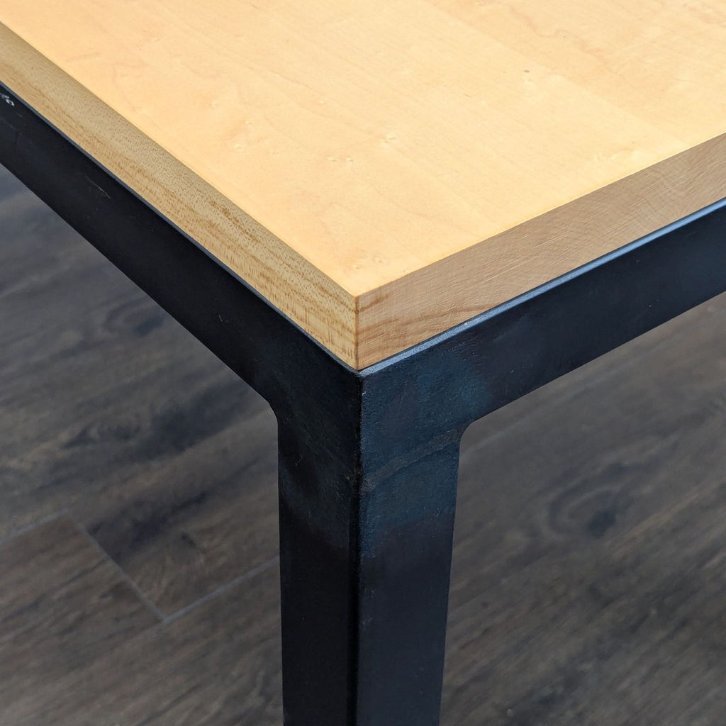 Close-up of a Room & Board maple dining table corner, highlighting the natural steel base and wood grain.