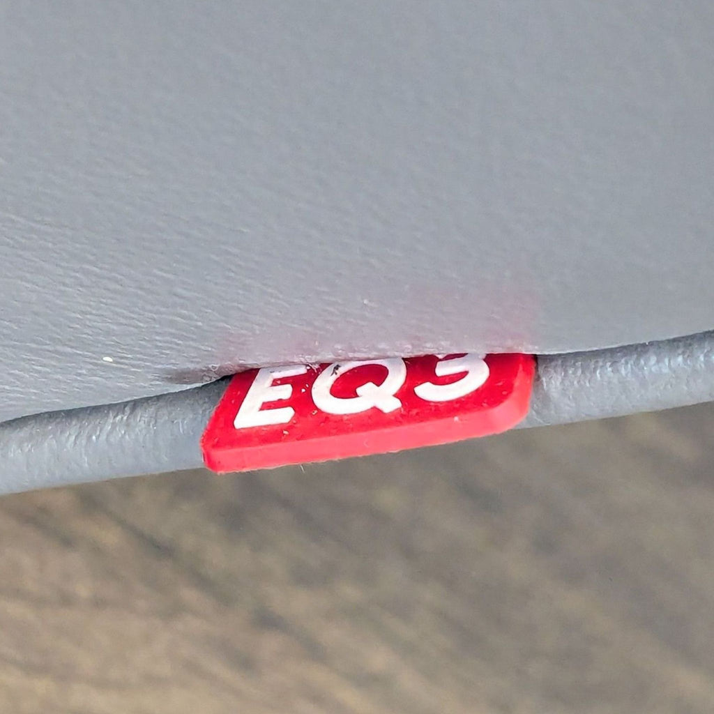 2. Red EQ3 brand tag attached to the seam of grey leather upholstery, seen up close.