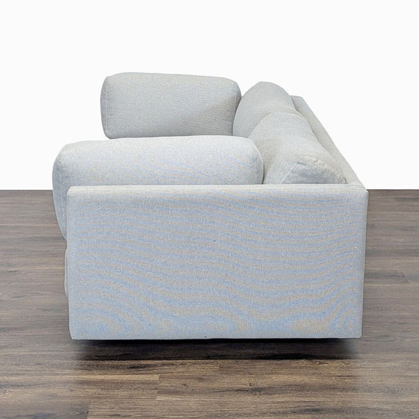 1. Side view of a BluDot 3-seat sofa in light fabric on a wooden floor, showing the armrest and cushions.