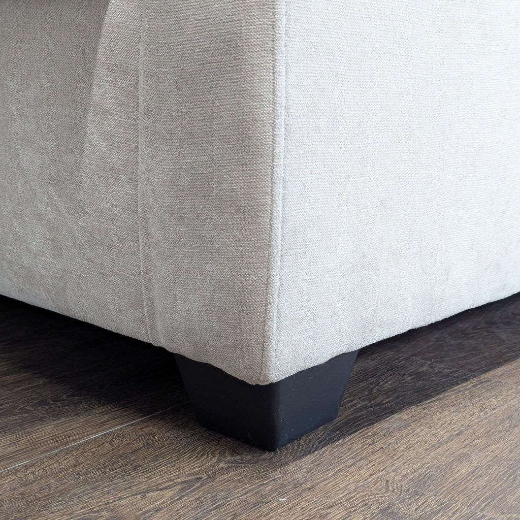Detail of a Living Spaces 3-seat sofa corner showing light beige fabric and black wooden leg on wood flooring.  