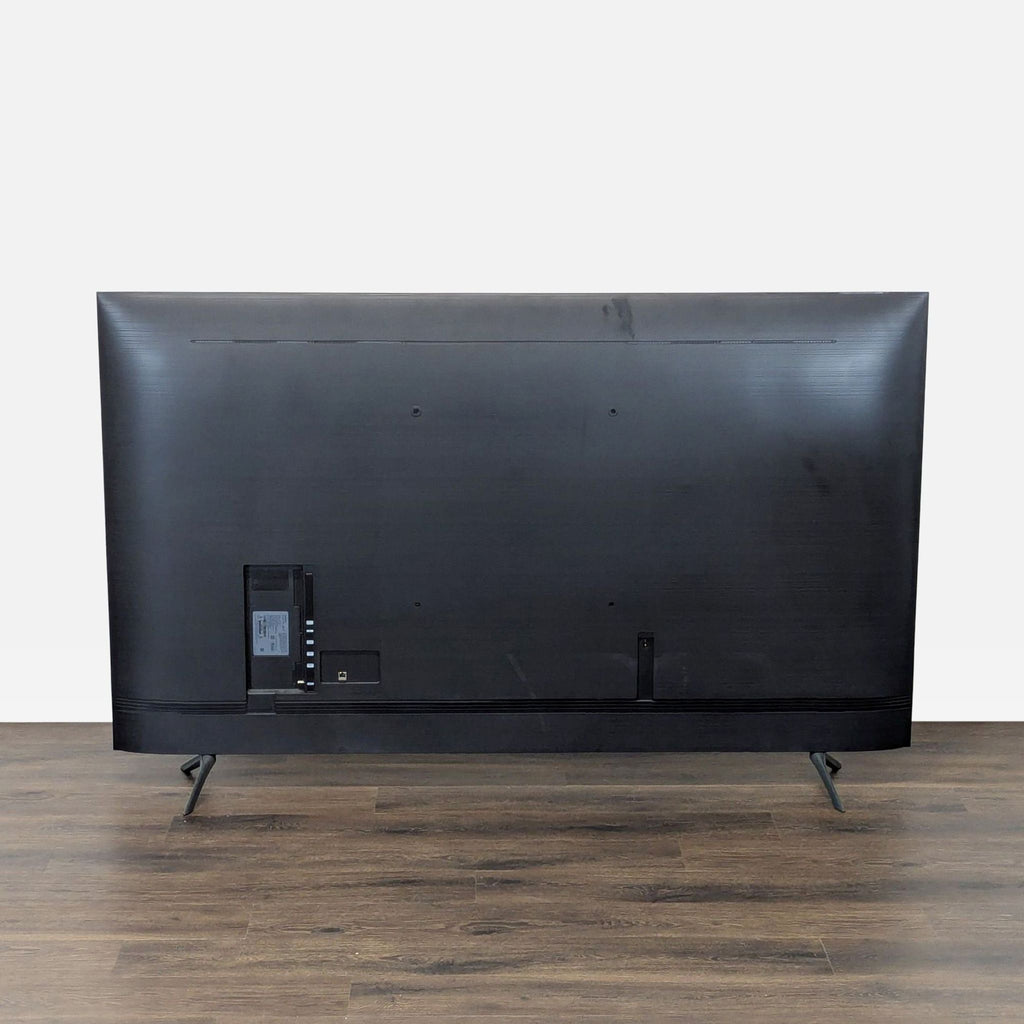 Back view of a Samsung TV showing its black textured surface and stand, placed on a wooden floor.  
