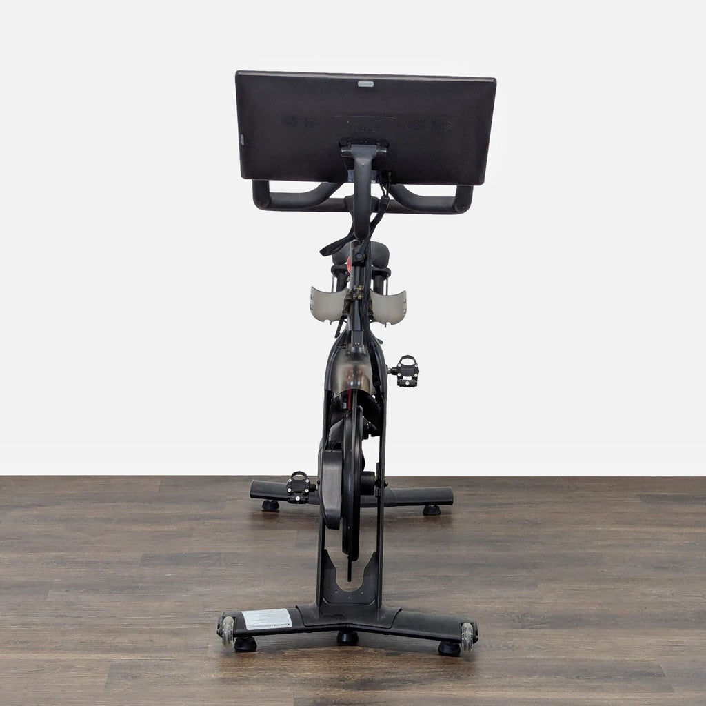 3. Rear view of a Peloton gym bike highlighting the frame, screen, and pedals on a wood surface.