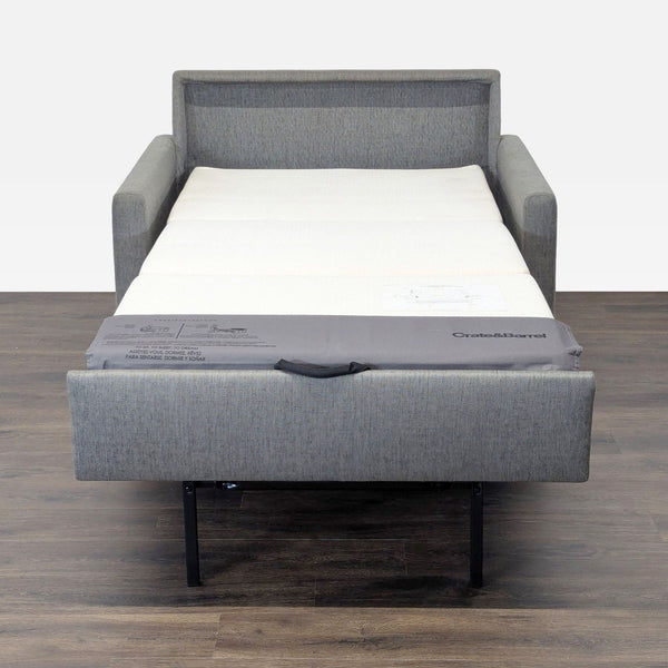 Gray Crate & Barrel sleeper sofa fully extended into a bed, shown against a wood floor and plain wall.  