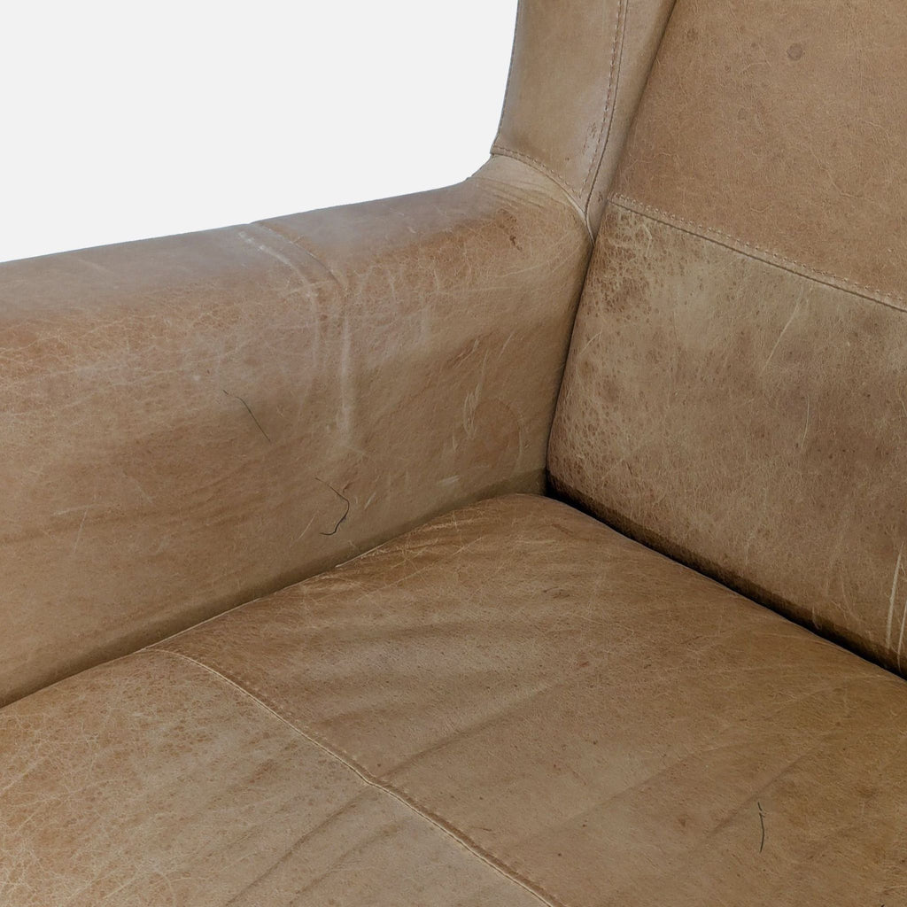 2. Close-up of the worn tan leather seat and arm of a West Elm lounge chair, highlighting visible texture and scratches.