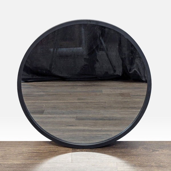 Round Umbra mirror with a black frame, reflecting wood flooring and a dark curtain in the background.  