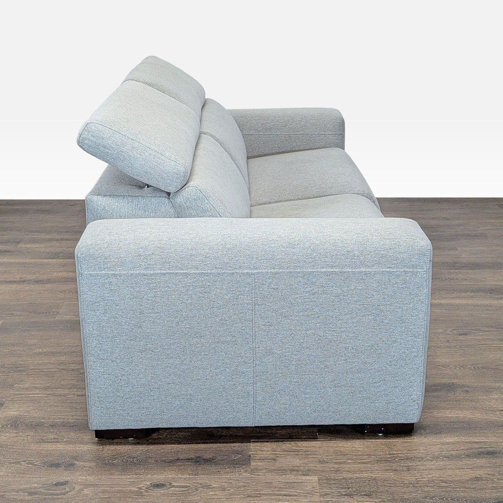 3. Side view of a light gray Macy’s sofa with thick armrests and adjustable headrests on a wood floor.