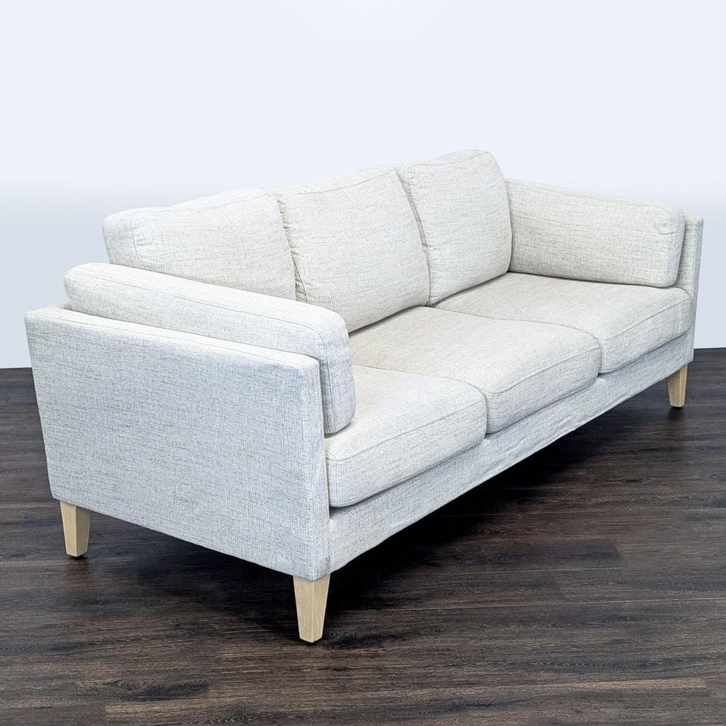3. Angled view of a light gray 3-seat World Market sofa with thick cushions and natural wood legs on a dark floor.
