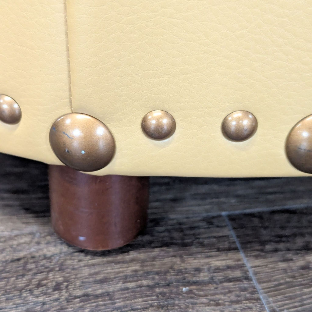3. Close-up of Reperch ottoman leg, featuring brown wood and detailed brass nailhead accents on tan leather.