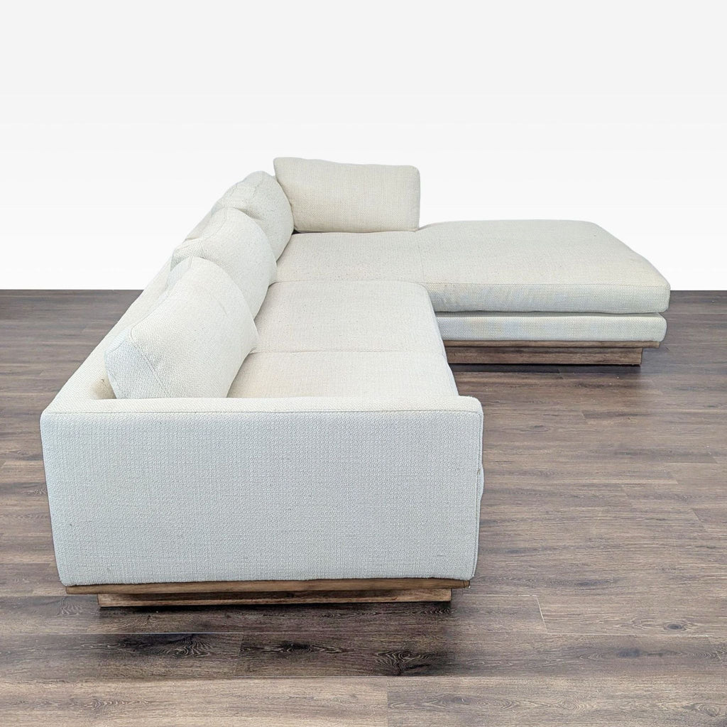 2. Side view of a modern beige Reperch sectional with firm back cushions and wooden base on dark flooring.
