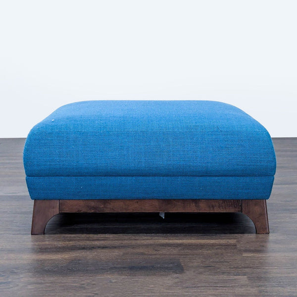 1. Blue upholstered ottoman by Article with wooden legs, shown in front view on a dark wooden floor.