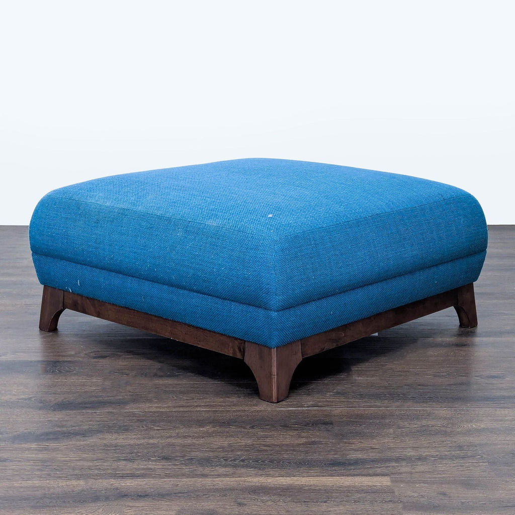 1. Square blue ottoman by Article with wooden legs, shown on dark wood flooring from an angled view.  