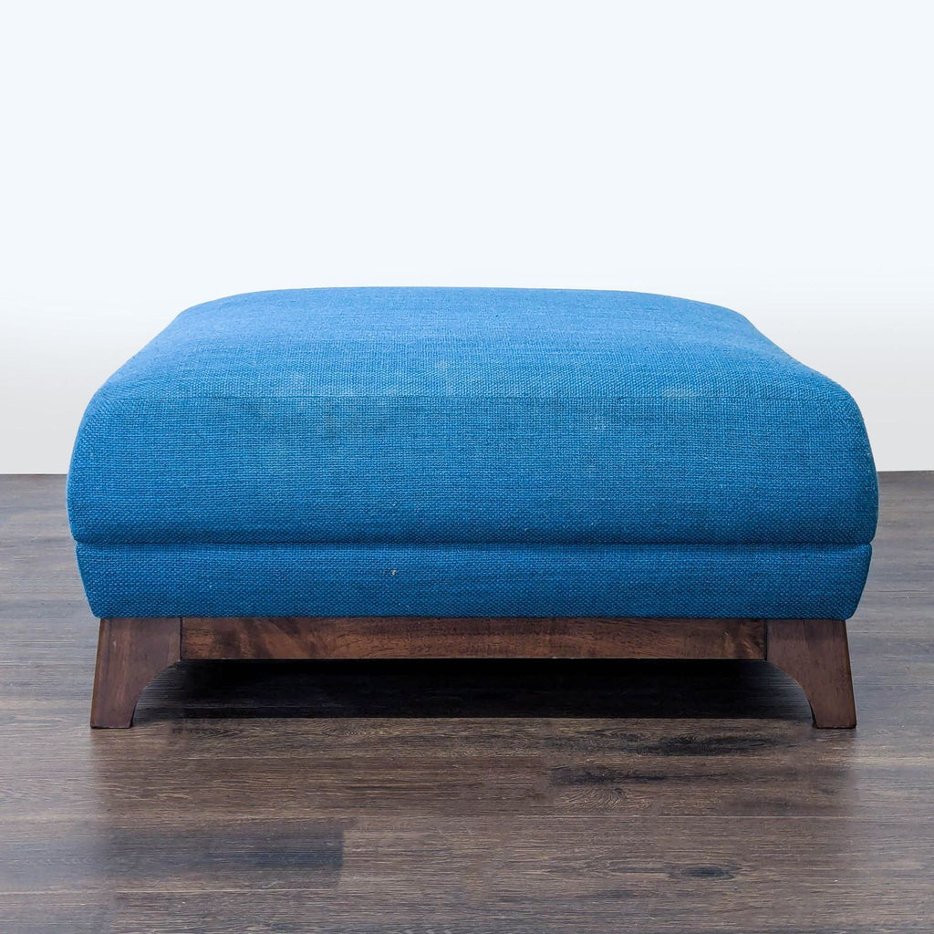 3. Blue Article ottoman with wooden legs, displayed from a direct front view on dark wood floor.