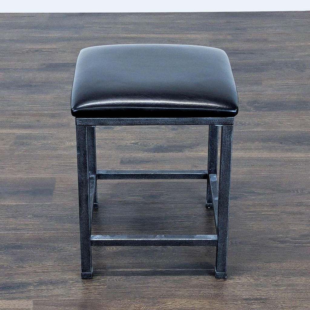2. Overhead view of a black padded square stool with a metal base, positioned on a wooden floor.  
