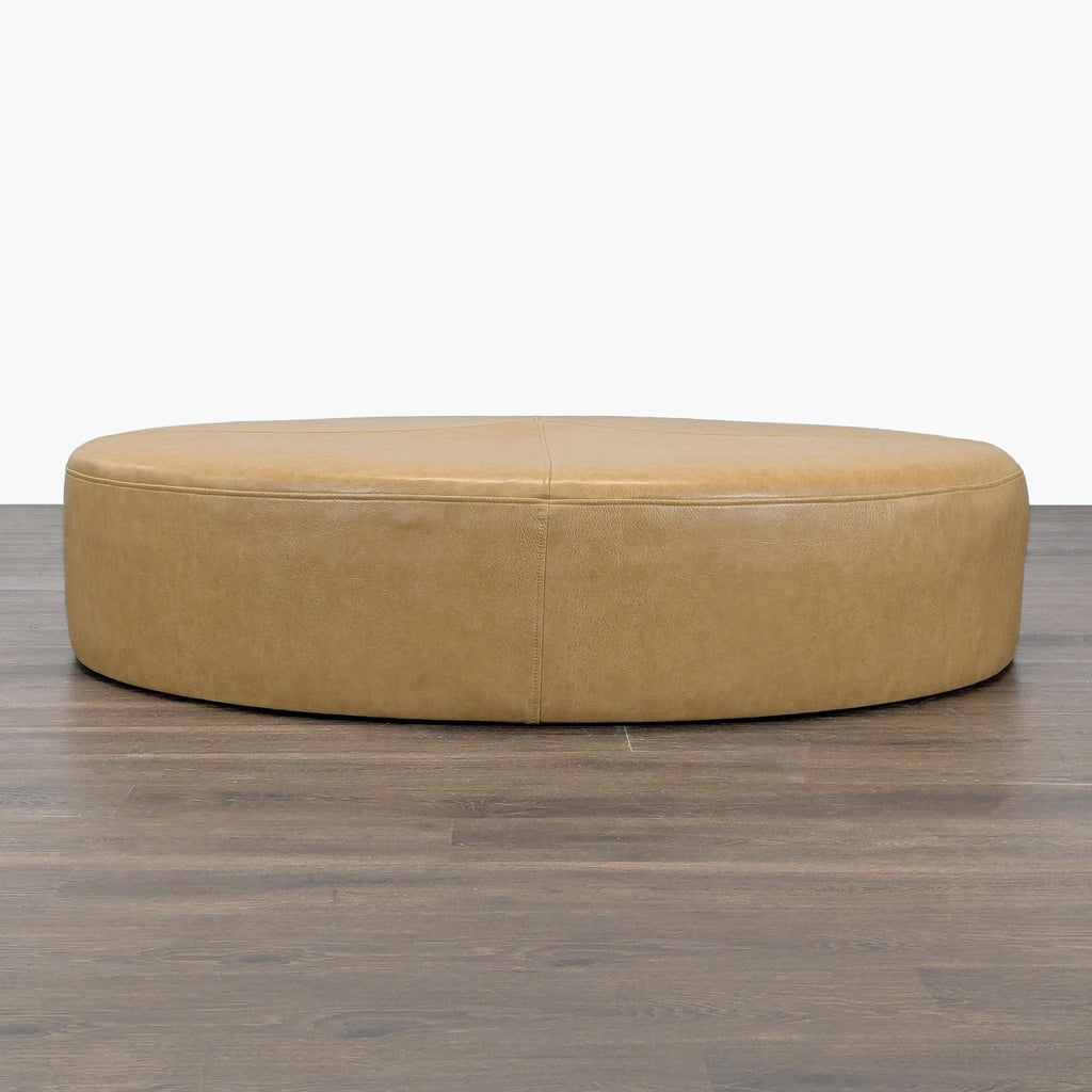 1. Large round tan ottoman with smooth faux leather upholstery, shown from a front angle on wood flooring.  