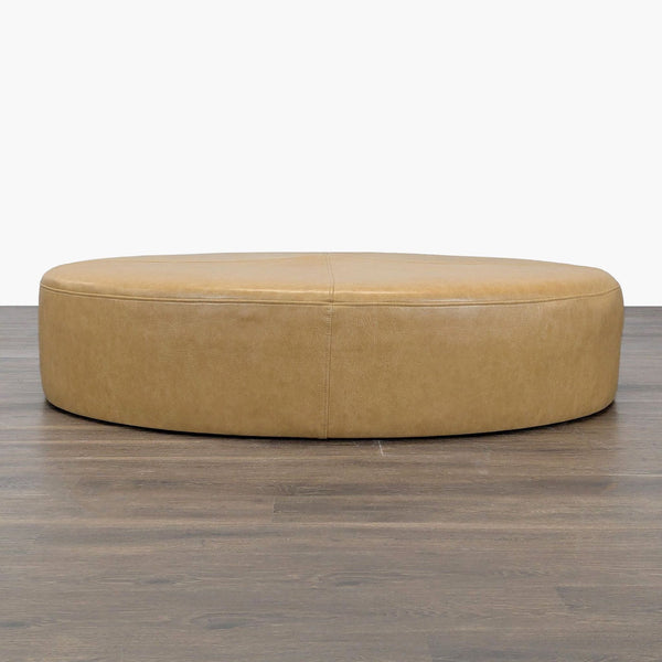1. Large round tan ottoman with smooth faux leather upholstery, shown from a front angle on wood flooring.  