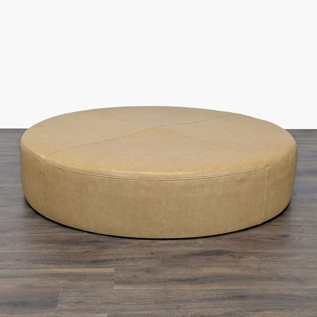 2. Wide circular tan ottoman, featuring subtle stitching details, sits on a wooden floor with a plain backdrop.  