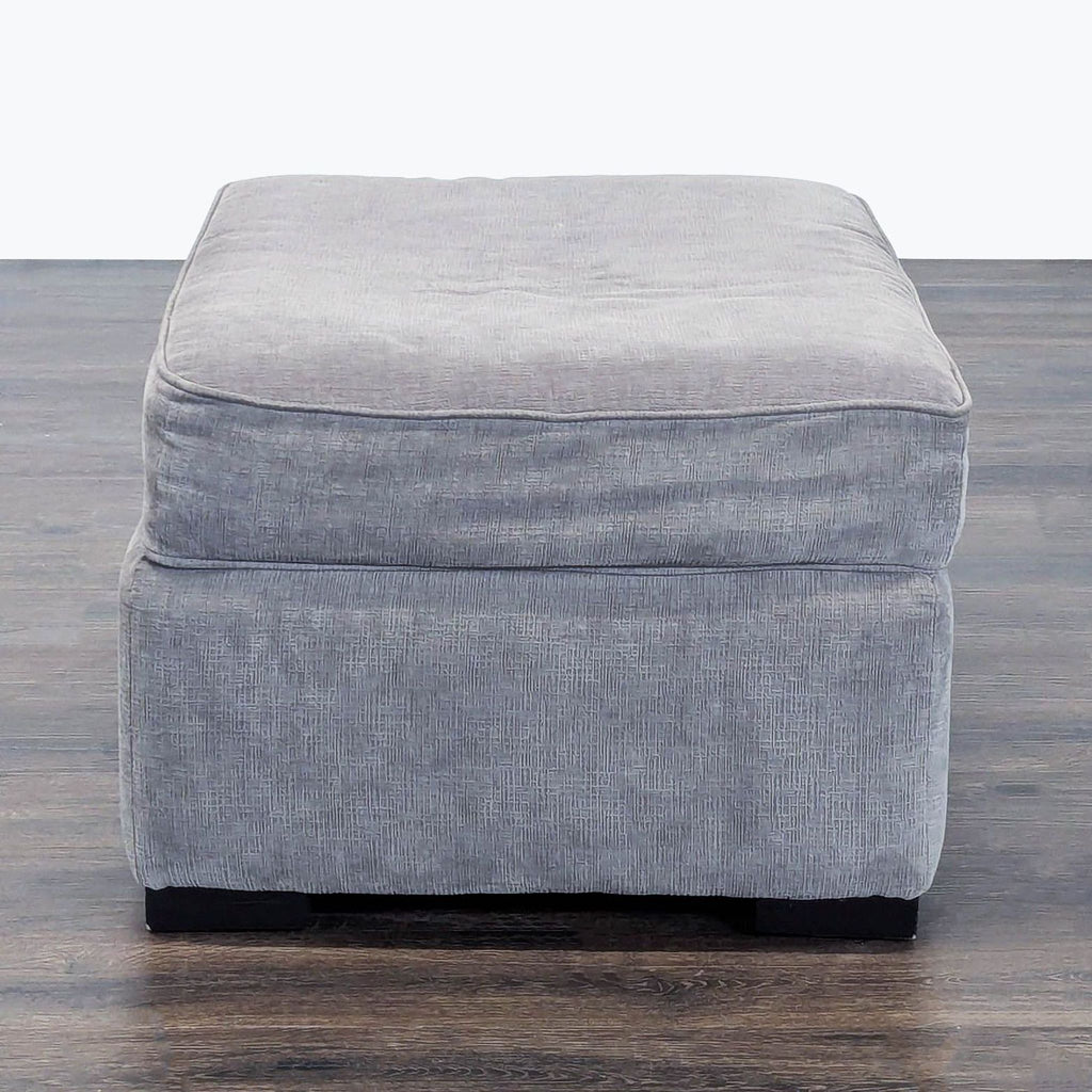 3. Side perspective of a light gray cushioned ottoman with block legs on a wooden floor.