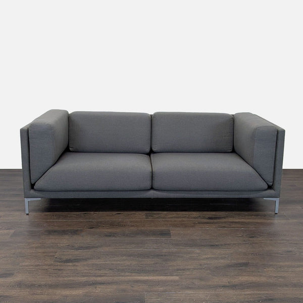 CB2 loveseat with dark gray upholstery, boxy shape, deep cushions, and metal legs, ideal for modern spaces.