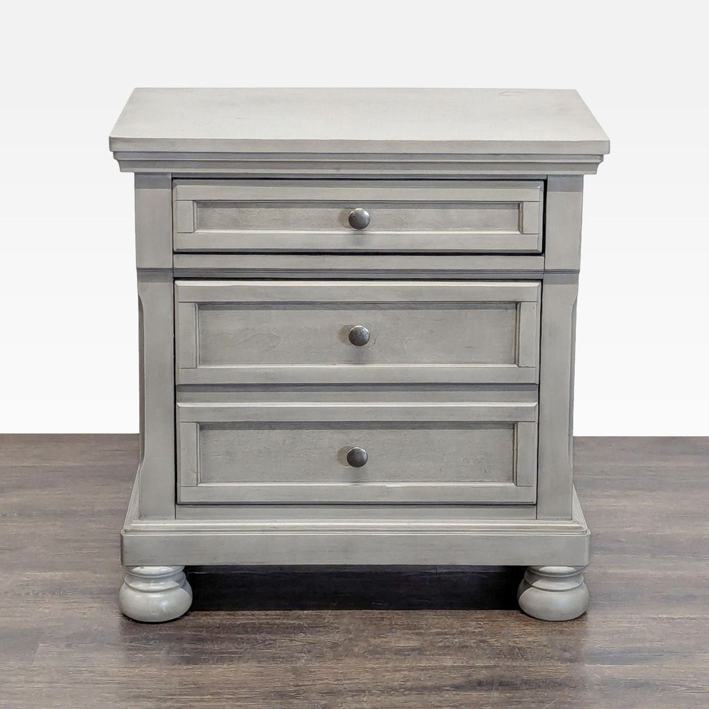 Light gray end table by Ashley Furniture with two drawers, bun feet, inlaid panels, and silver hardware.