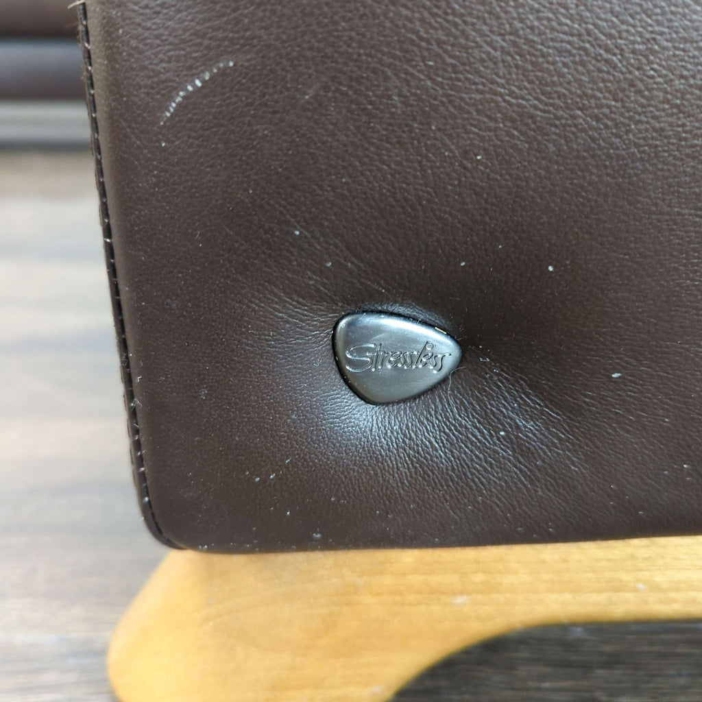 3. Detailed view of Stressless brand logo on brown leather with visible stitching and wood leg base.