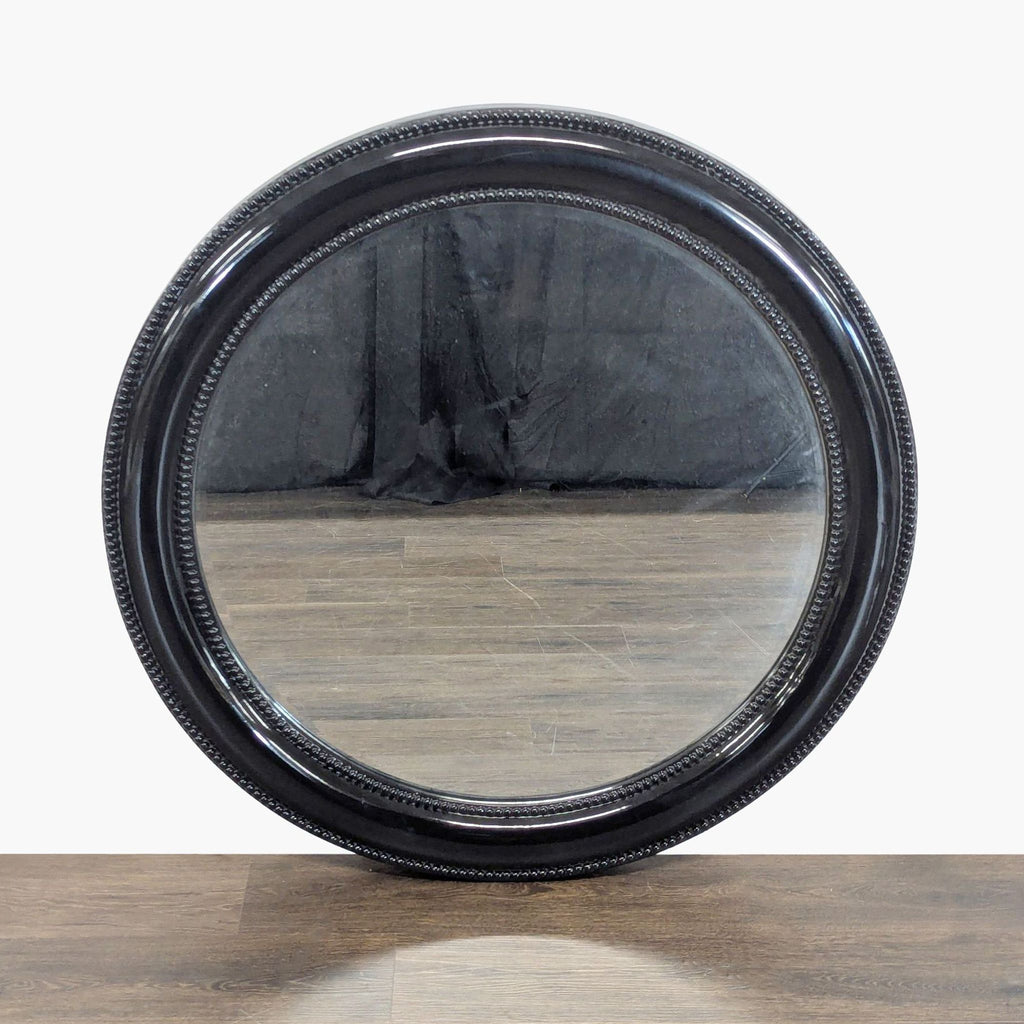Round Howard Elliott mirror with a black resin frame, shown upright on a wood surface.  