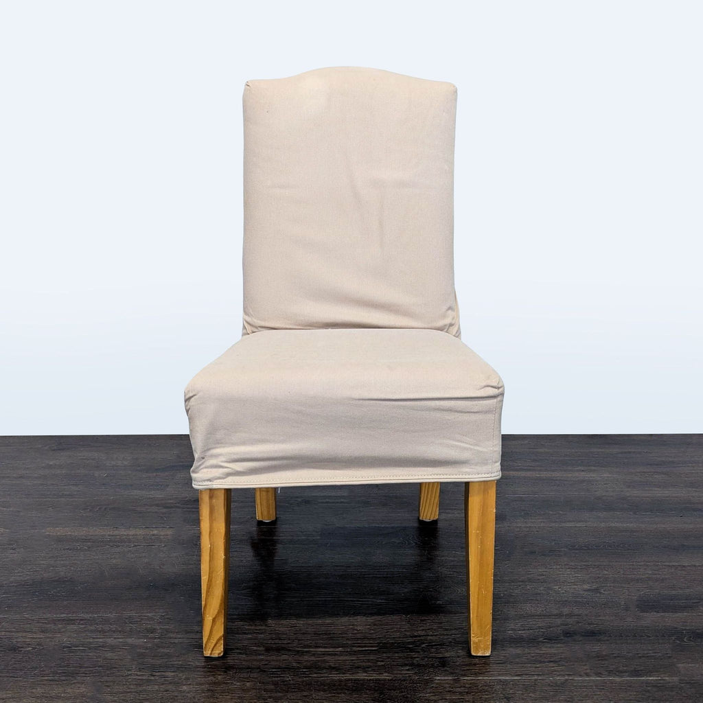 2. Single beige slipcovered dining chair with wooden legs, shown front-facing on dark wood floor.  