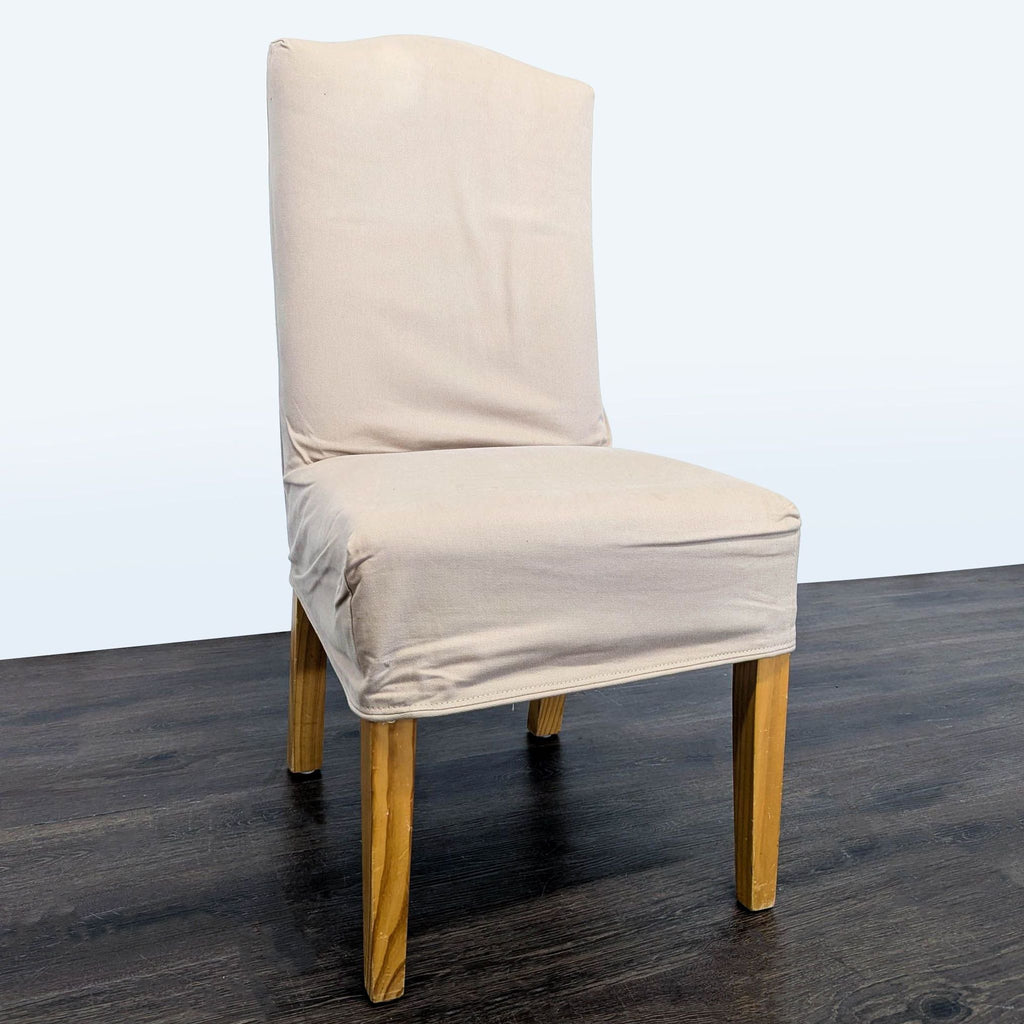 3. Angled side view of a beige slipcovered dining chair with wooden legs on dark wood flooring.