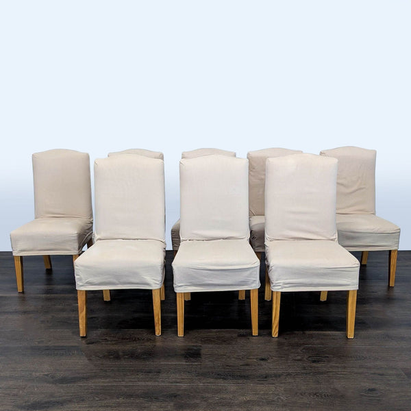 1. Set of eight beige slipcovered dining chairs with wooden legs arranged on dark wood flooring.  