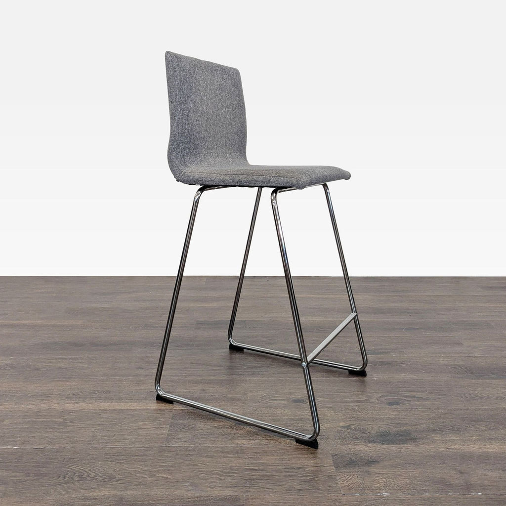 Modern grey upholstered counter stool with sleek metal legs by Ikea, photographed on a dark wood floor.