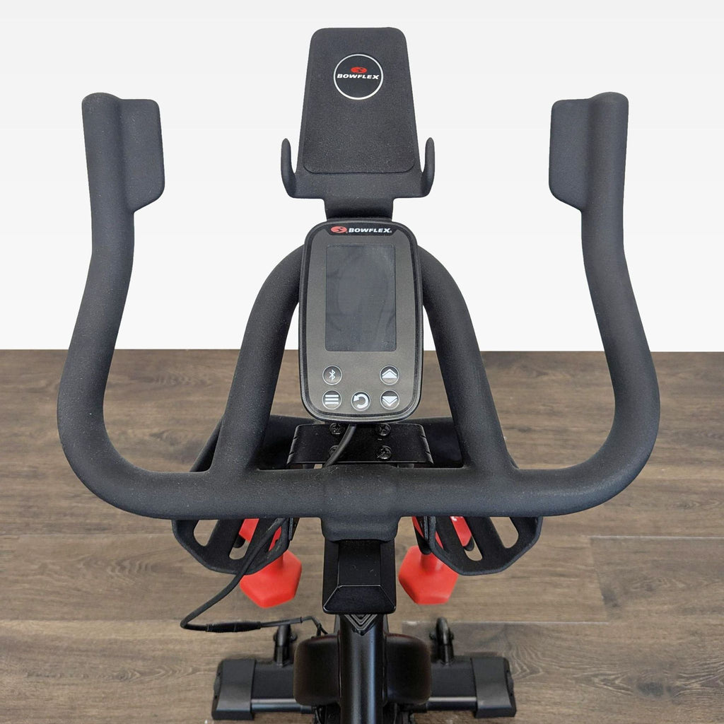 Close-up of Bowflex C6 bike handlebars with digital display and storage for two red dumbbells underneath.