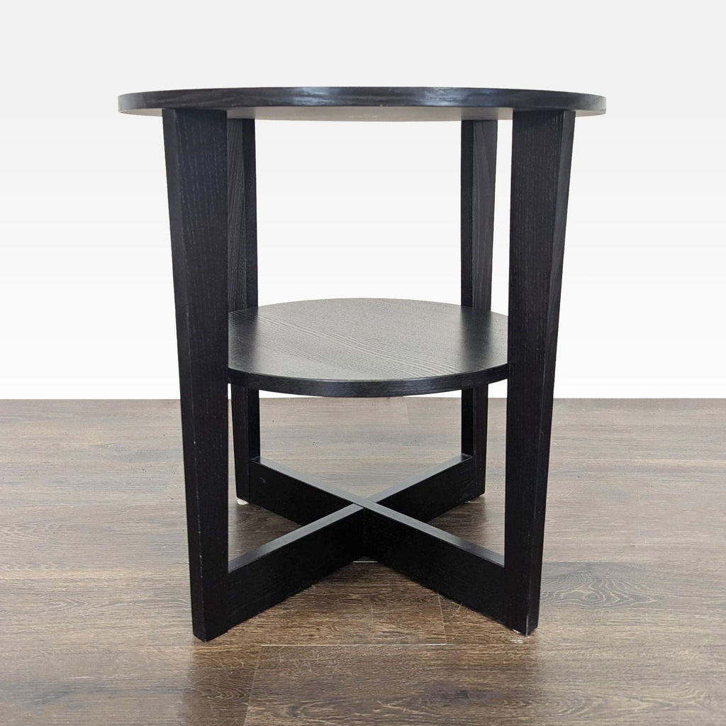 3. Front view of a black round end table featuring a lower shelf and X-shaped base, on hardwood floor.