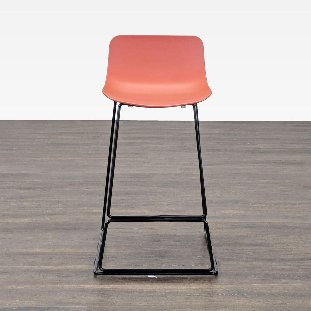 2. Front view of Article bar stool featuring a coral seat and sleek black metal legs on wooden floor.