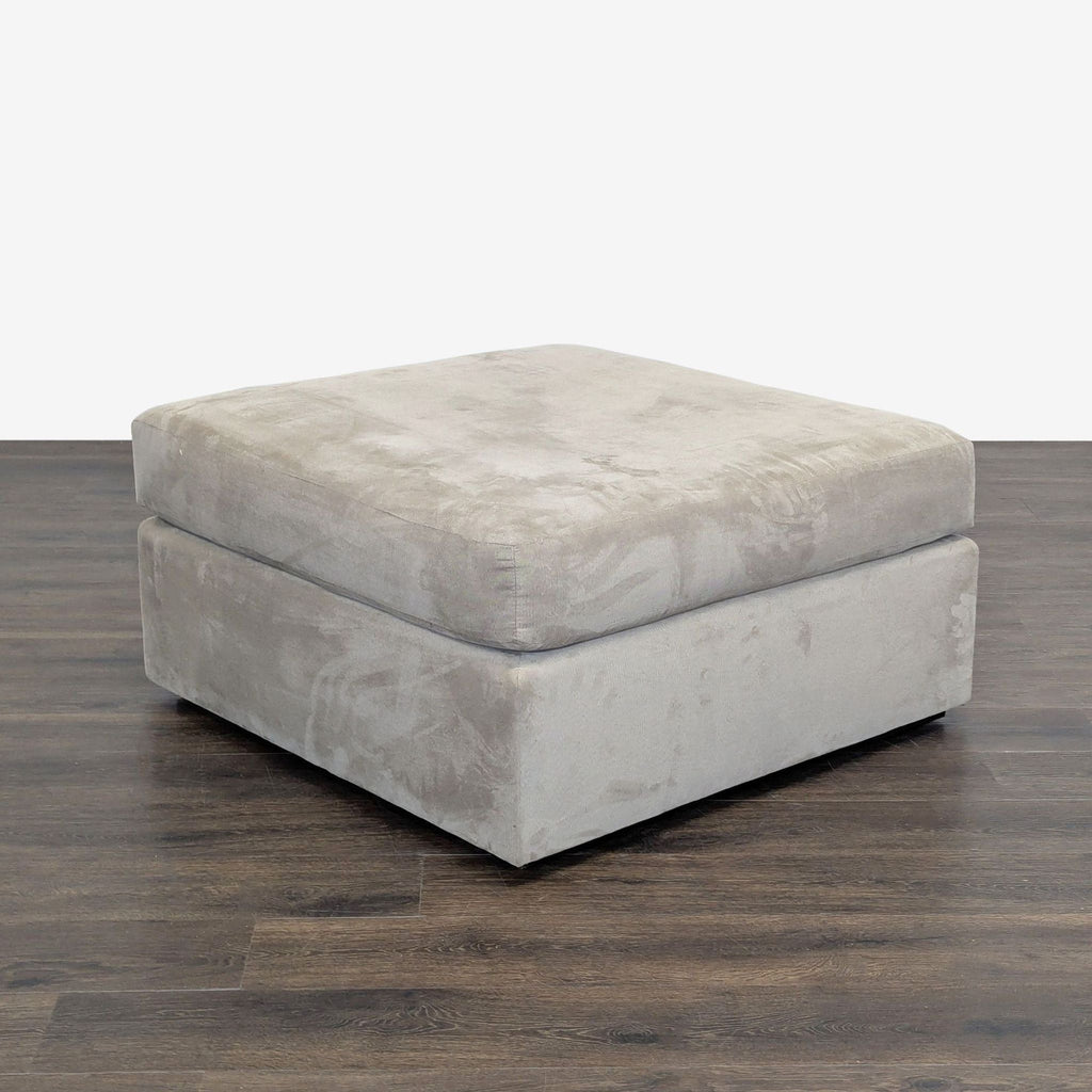 2. Angled view of a light grey, soft-cushioned ottoman with a low profile, on a wooden floor.
