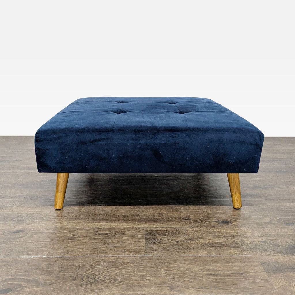 3. Front view of a square, tufted navy blue ottoman with wooden legs, placed on a wood floor.