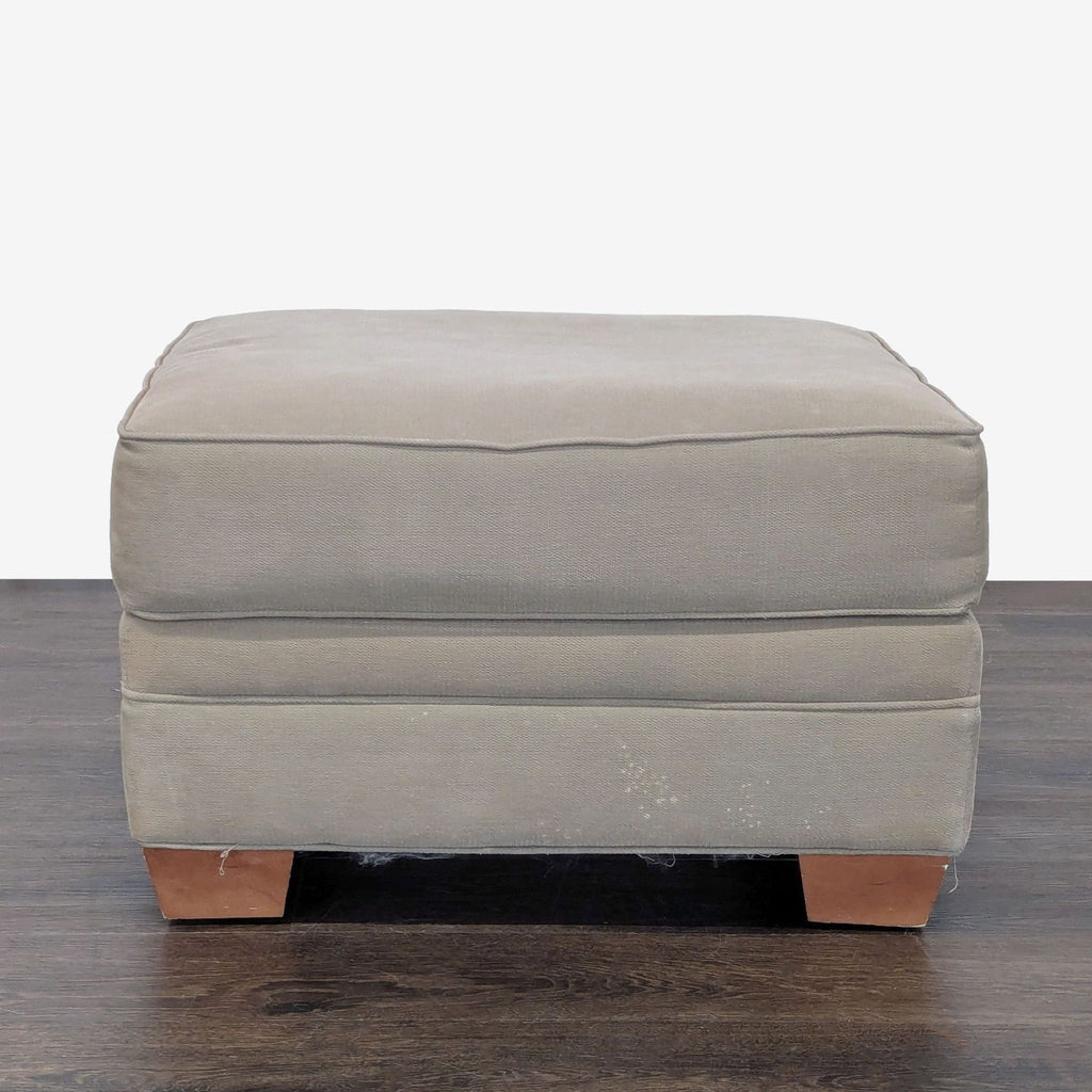 2. Front view of a gray upholstered ottoman with wooden block feet on a dark hardwood surface.