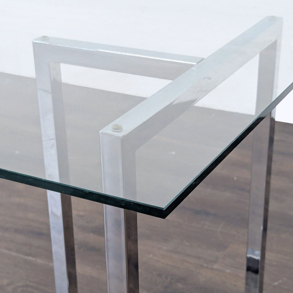 Close-up of a CB2 dining table with a clear glass top and chrome-finished metal frame on a wood floor.  