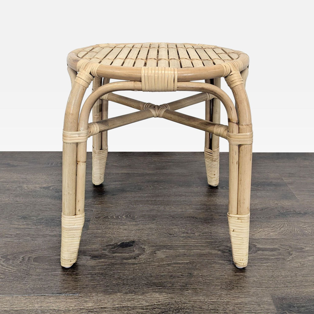 Single round rattan stool, featuring a woven top and sturdy legs, shown on a dark wooden floor.  